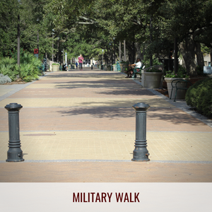 Looking down the brick paved Military Walk of Texas A&M campus. Large oak trees shade a path that travels between campus buildings and the main academic building and courtyard.